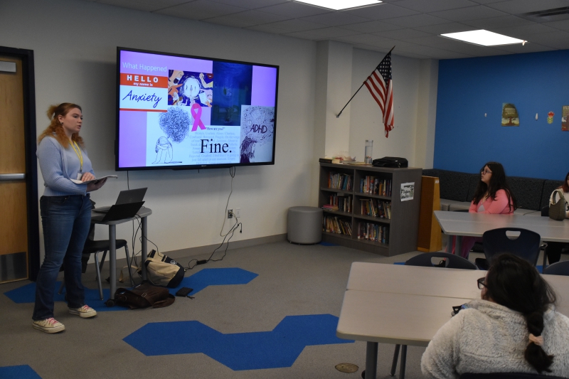 Makayla Powder, a NAMI young Adult presenter, shares her story with Resilience Academy students. A background slide shows a nametag with 'Hello my name is Anxiety,' an illustration of people screaming at someone, someone with a swirl of multiple things and ADHD and another a girl with a tangled ball of confusion above her